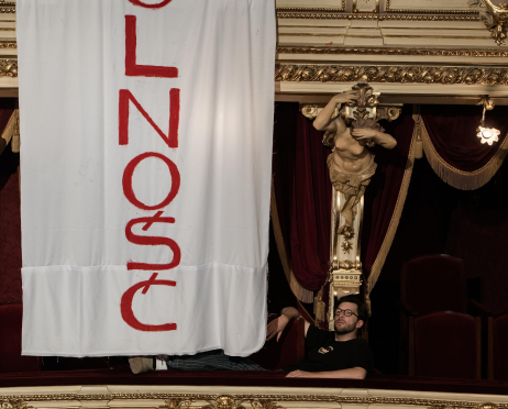 Balkon w bogato zdobionym wnętrzu teatru. Z balustrady zwisa duży biały transparent z czerwonym, pionowym napisem „WOLNOŚĆ”. Obok niego, w loży, siedzi aktor Mateusz Bieryt ubrany na czarno, oparty swobodnie o poręcz.